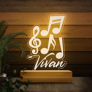 May include: A lighted acrylic music-themed night light. The design features musical notes and the name "Vivan" in a white, illuminated script. It sits on a rectangular wooden base, creating a warm, ambient glow. The background is a wooden wall.