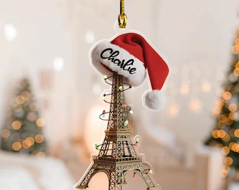 May include: A Christmas ornament featuring the Eiffel Tower decorated with string lights and topped with a Santa hat. The hat is red with white trim and the name "Charlie" is written on it. The ornament hangs from a gold string.