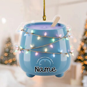 May include: A blue wax warmer ornament with a string of colorful lights wrapped around it. The warmer has a dial and the word "Name" in black script. A wooden stick rests in the purple wax.