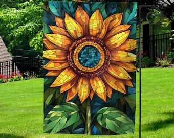 May include: A decorative garden flag featuring a stained-glass style sunflower design. The sunflower has yellow and orange petals, a blue center, and green leaves. The flag is displayed on a black metal stand in a grassy yard.