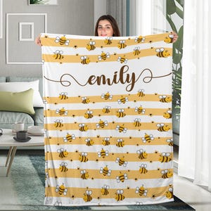 May include: A white and yellow striped blanket featuring cartoon bees and the name "emily" in brown script. The blanket has a repeating pattern of bees and yellow stripes, creating a cozy and cheerful design.