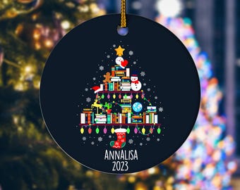 May include: A round, dark blue Christmas ornament with a gold string. The ornament features a Christmas tree design made of books, a snowman, and a stocking. The name "Annalisa" and the year "2023" are printed on the bottom.