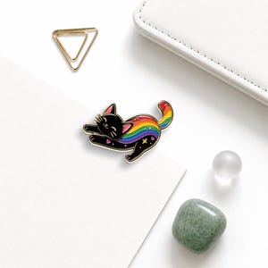May include: A black enamel pin shaped like a stretching cat with a rainbow-colored back, gold-tone metal outlines, and small star and heart accents. The cat has closed eyes, a smiling mouth, and whiskers, with the rainbow pattern extending to its tail.