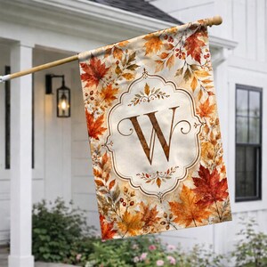 May include: A rectangular garden flag featuring a pattern of autumn leaves in shades of orange, red, and yellow. In the center, a decorative white frame with a scalloped border encloses a large, dark brown capital letter W in a serif font.