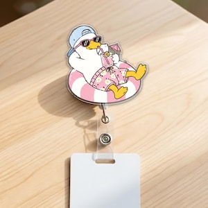 May include: A badge reel featuring a white duck wearing a blue baseball cap and black sunglasses, lounging in a pink and white striped inner tube. The duck wears pink floral shorts and holds a pink drink with a small umbrella and a straw.