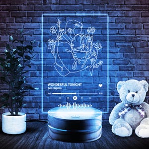 May include: A blue neon sign with a line drawing of a couple embracing, surrounded by flowers. The sign has a music player interface with the song "Wonderful Tonight" by Eric Clapton playing. The sign is on a wooden base.