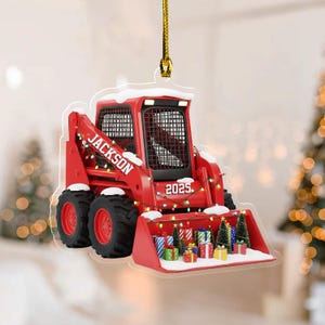 May include: A red skid steer loader Christmas ornament with the name "Jackson" and the year "2025" printed on it. The ornament is decorated with Christmas lights, snow, and miniature presents and trees.