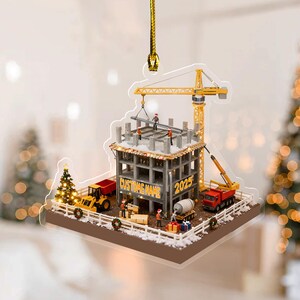 May include: A clear acrylic Christmas ornament depicting a construction site with a building under construction, a crane, and construction vehicles. The ornament has the text "CUSTOM NAME" and "2025". The ornament is hanging from a gold string.