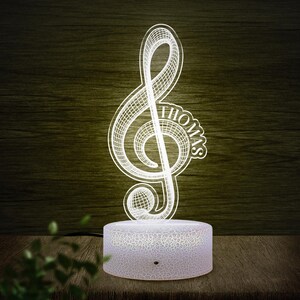 May include: A white, illuminated treble clef night light with the name "THOMAS" etched into the design. The light sits on a round, textured base. The background is a dark wood grain.