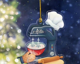 May include: A clear acrylic ornament featuring a blue stand mixer, a red bowl with whipped cream, a chef's hat, a rolling pin, and a whisk. The name "Alita" is written on the mixer. The ornament is suspended by a gold string.