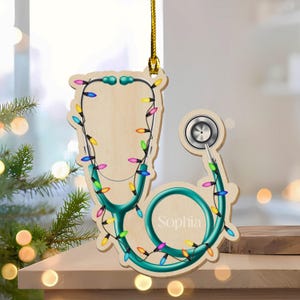 May include: Wooden stethoscope Christmas ornament with teal accents and colorful lights. The name "Sophia" is printed on the stethoscope. A gold string is attached for hanging. The ornament is on a wooden surface.