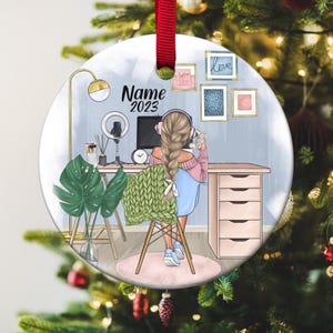 May include: Round Christmas ornament with a red ribbon. The ornament features an illustration of a person at a desk with a computer, headphones, and a microphone. The text "Name 2023" is above the illustration. Decorative art is on the wall.