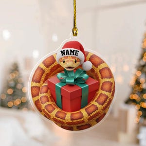 May include: A Christmas ornament featuring a cartoon snake wearing a Santa hat with the word "NAME" on it. The snake is coiled around a red gift box with a green bow. The ornament is suspended by a gold string.