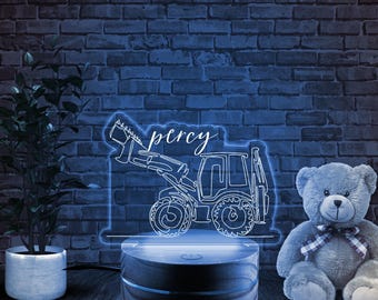 May include: A white outline of a tractor with the name "Percy" written above it, illuminated by a blue light. The tractor is on a wooden base with a blue light underneath. A gray teddy bear is sitting to the right of the tractor.