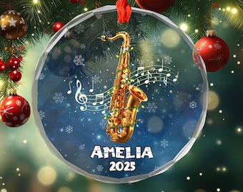 May include: A clear, round Christmas ornament featuring a gold saxophone decorated with lights and musical notes. The ornament has the name "AMELIA" and the year "2025" printed on it. The background is a blue gradient with snowflakes.