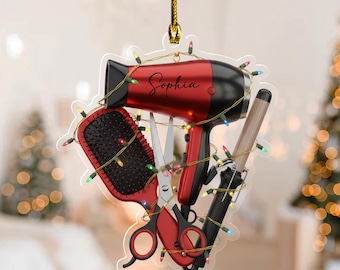 May include: A clear ornament featuring a red and black hair dryer, hairbrush, scissors, and curling iron, all adorned with a string of colorful Christmas lights. The name "Sophia" is written on the hair dryer. A gold string is attached for hanging.