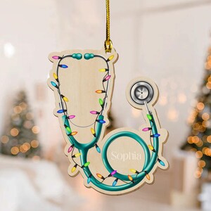 May include: Wooden stethoscope Christmas ornament with teal accents and colorful lights. The ornament has the name "Sophia" on it. The ornament is hanging from a gold string.