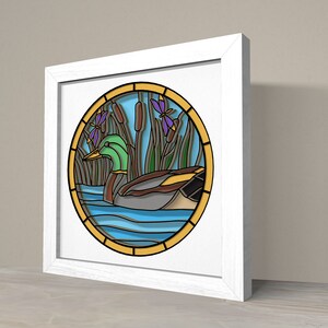 May include: A framed stained glass artwork depicting a green duck swimming in blue water with yellow cattails and purple flowers.