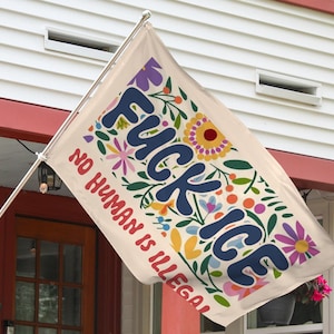 May include: A rectangular flag with the words "FUCK ICE" in navy blue, surrounded by colorful floral designs. The phrase "NO HUMAN IS ILLEGAL" is in red. The flag is hanging outside a house.