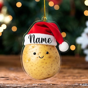 May include: A clear acrylic Christmas ornament shaped like a potato with a smiling face and a red Santa hat. The word "Name" is written in black script on the hat. The ornament hangs from a gold string.