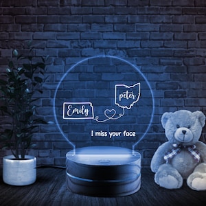 May include: A personalized LED night light with a circular design. The light features the names "Emily" and "Peter" with a heart connecting them, and the text "I miss your face." The base is a dark wood color.