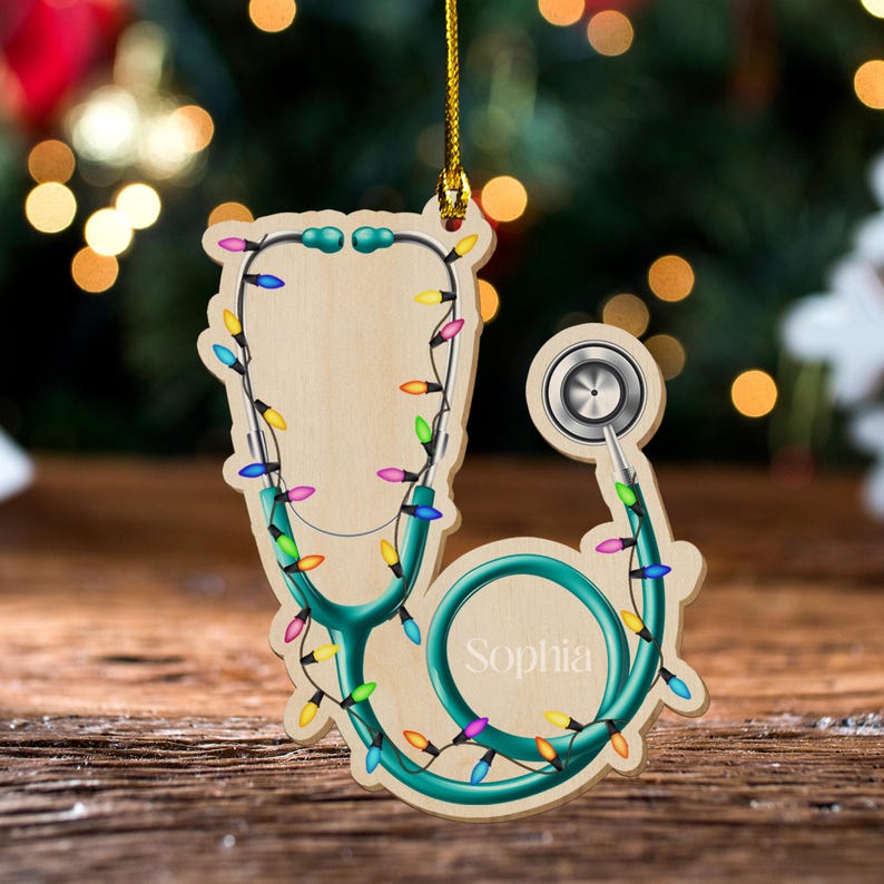 May include: Wooden stethoscope Christmas ornament with teal accents and colorful lights. The ornament has the name "Sophia" on it. The ornament is hanging from a gold string.