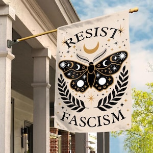 May include: A beige house flag with the words "RESIST FASCISM" and a black and gold moth design. The moth has crescent moon and star details, with a laurel wreath at the bottom. The flag is hanging on a gold pole.