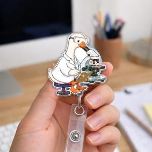 May include: An acrylic badge reel featuring a cartoon white duck wearing a lab coat and safety goggles, looking through a microscope while holding a clipboard. The design includes sparkling glitter accents on the clipboard and the microscope base.