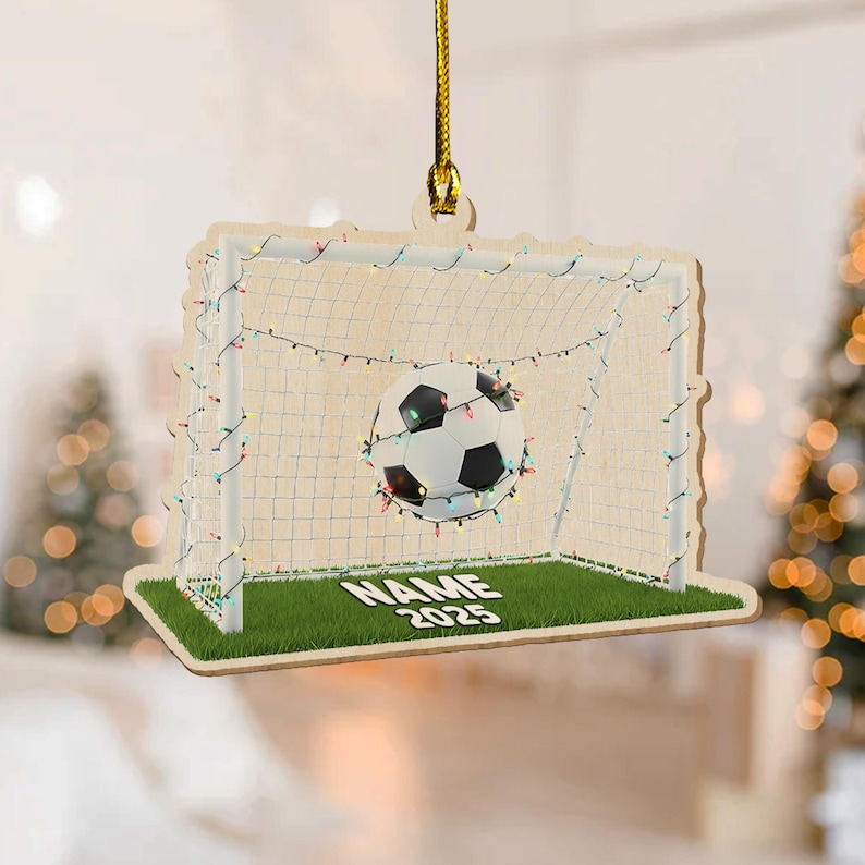 May include: Wooden soccer-themed ornament featuring a soccer ball in a net, decorated with string lights. The ornament has a green field base with the text "NAME 2025". The ornament hangs from a gold string.