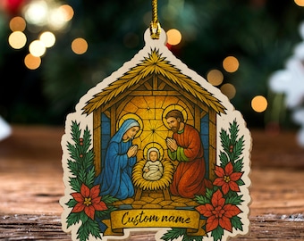 May include: A Christmas ornament featuring a Nativity scene with Mary, Joseph, and baby Jesus. The ornament is shaped like a stable, with a gold string for hanging. The ornament has a banner that says "Custom name" and is decorated with red poinsettias.