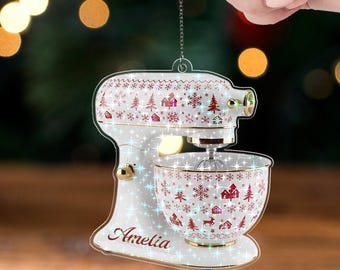May include: A white and gold acrylic Christmas ornament shaped like a stand mixer. The ornament is decorated with red and white snowflakes, trees, and houses. The name "Amelia" is written on the base. The text "ACRYLIC - TWINKLE" is at the bottom.