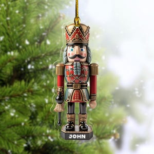 May include: A Christmas ornament shaped like a nutcracker soldier. The ornament is red, blue, and gold with the name "JOHN" at the base. It hangs from a gold string against a blurred Christmas tree background.