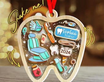 May include: Wooden ornament in the shape of a tooth, featuring dental tools and a dental chair. The ornament has the text "Sophia Nurse" and "2024". A red ribbon is attached for hanging. The words "Shake me!" are written on the ornament.