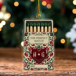 May include: A decorative ornament shaped like a matchbox, featuring the text "THE PERFECT MATCH" and a heart with the initials "A + G". The match heads are green, and the box has a floral design in red and green.
