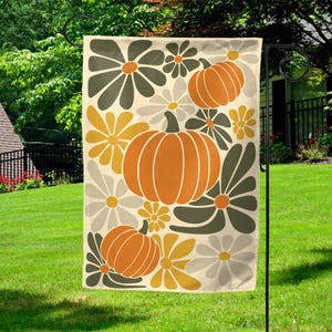 May include: A decorative garden flag featuring a repeating pattern of orange pumpkins and stylized flowers in shades of yellow, gray, and olive green on a cream background. The flag is displayed on a black metal stand in a grassy yard.