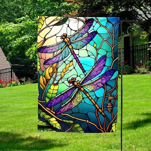 May include: A decorative garden flag featuring a stained-glass style design with two dragonflies. The dragonflies have purple and yellow wings and are set against a backdrop of blue, green, and yellow. The flag is displayed on a black metal stand.