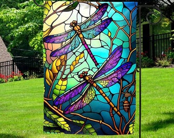 May include: A decorative garden flag featuring a stained-glass style design with two dragonflies. The dragonflies have purple and yellow wings and are set against a backdrop of blue, green, and yellow. The flag is displayed on a black metal stand.