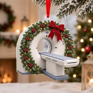 May include: A white CT scanner ornament featuring a festive green holly wreath with red berries and small warm lights around the circular frame. A large red ribbon bow sits at the top. The scanner bed is labeled with the text LUCAS-202X in black.