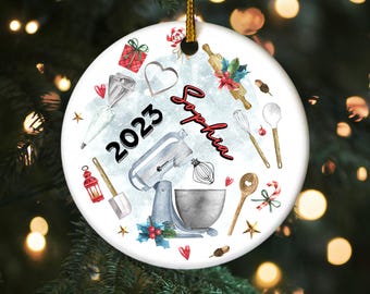 May include: Round ceramic Christmas ornament with a gold string. The ornament features watercolor illustrations of baking tools, a stand mixer, and the text "2023" and "Sophia". The background is light blue with small stars and hearts.