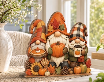 May include: A flat, decorative wooden cutout featuring three gnomes with long white beards. The gnomes wear orange, plaid, and buffalo check hats adorned with autumn leaves. They hold a pumpkin pie, a large orange pumpkin, and a coffee mug.