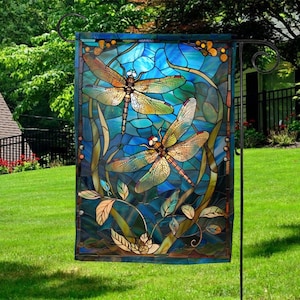 May include: A decorative garden flag featuring a stained-glass design with two dragonflies. The dragonflies have orange and yellow wings, set against a blue background with green and brown accents. The flag is displayed on a black metal stand.
