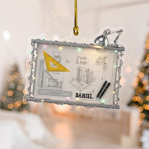 May include: A rectangular ornament featuring an architectural blueprint design with a yellow triangle ruler, a silver desk lamp, and the name "DANIEL". The ornament is framed with a string of colorful lights and suspended by a gold cord.