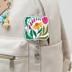 May include: A floral badge reel featuring a colorful design of a yellow daisy, pink tulip, and bell-shaped flowers with green leaves. The badge reel is attached to a light-colored backpack with a retractable cord and a metal clip.