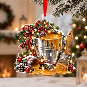 May include: A Christmas ornament featuring a silver paint bucket with the name JACK printed on it, a wooden paintbrush with 2026 printed on the handle, and golden paint spilling out. It is decorated with a plaid bow, pine branches, and red ornaments.
