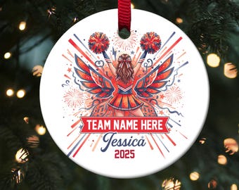 May include: A white round ornament with a cheerleader design. The ornament features a cheerleader with red and blue pom-poms, wings, and the text "TEAM NAME HERE", "Jessica", and "2025". A red ribbon is attached for hanging.