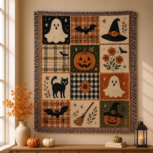 May include: A woven throw blanket featuring a grid of twelve squares with Halloween imagery including ghosts, a jack-o'-lantern, a black cat, a witch hat, a broom, bats, and floral patterns in orange, black, white, and green with fringed edges.