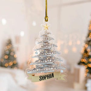 May include: A clear, Christmas tree-shaped ornament featuring a design of silver flutes. The ornament is topped with a gold star and has a banner with the name "SOPHIA". It hangs from a gold cord.