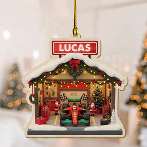 May include: Wooden Christmas ornament shaped like a garage with a race car inside. The ornament has a red sign that says "LUCAS". The garage is decorated with Christmas lights, a wreath, and a Christmas tree. Santa Claus figures are also present.