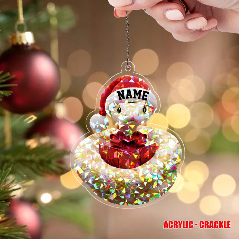May include: A clear acrylic Christmas decoration in the shape of a snake wearing a Santa hat. The ornament has a crackle effect and the word "NAME" is printed on the hat. A red gift box is in the center.