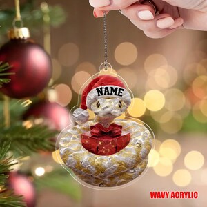 May include: A clear acrylic Christmas ornament depicting a snake wearing a Santa hat with the word "NAME" on it. The snake holds a red gift box with a bow. The ornament has gold and white details. The text "WAVY ACRYLIC" is at the bottom.